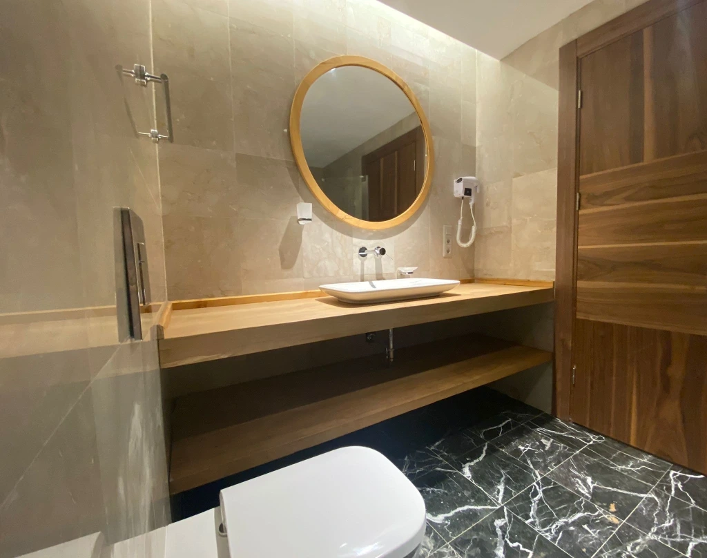 Yuvarlak aynalı ve ahşap lavabo tezgâhlı standart oda banyosu